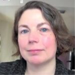 Portrait of a woman with short, wavy hair and blue eyes, wearing a dark sweater, set against a softly blurred background.