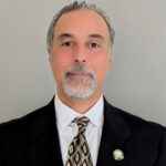 Man in a black suit with a patterned tie, sporting a goatee and a pin, stands against a light gray background.