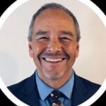 Smiling man with gray hair, wearing a blue suit and patterned tie, framed in a circular white border against a light background.