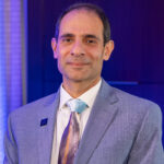 Man in a light blue suit with a patterned tie, sitting against a backdrop of soft purple lighting, smiling confidently.