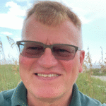 Man with glasses smiling, wearing a green polo shirt branded with "Geosynthetic Institute," set against a backdrop of beach grass and sky.