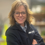 Smiling woman with shoulder-length, wavy hair wearing glasses and a dark zip-up jacket, arms crossed, against a blurred background.