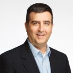 Smiling man with short dark hair wearing a dark jacket over a light blue checked shirt, set against a plain white background.