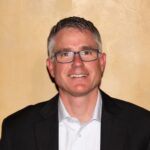 Smiling man with short hair and glasses, wearing a dark blazer over a light blue shirt, set against a warm, textured background.